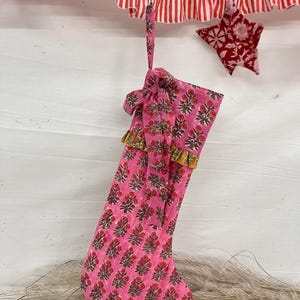 Könnte beinhalten: Ein rosa Weihnachtsstrumpf mit einem Blumenmuster aus roten Blüten und grünen Blättern. Der Strumpf hat eine goldene Verzierung und eine Schlaufe zum Aufhängen. Eine rot-weiß gestreifte Girlande und ein roter sternförmiger Schmuck sind im Hintergrund.