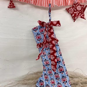 Puede incluir: Un calcetín navideño azul con un estampado floral, adornado con un lazo rojo y un lazo para colgar. Una guirnalda a rayas rojas y blancas y una decoración en forma de estrella completan la escena festiva.