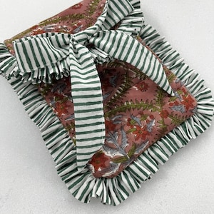 Puede incluir: Un cojín cuadrado con estampado floral en tonos rosa empolvado, rojo y gris, con borde con volantes y lazo a rayas verdes y blancas. El cojín es probablemente de tela.