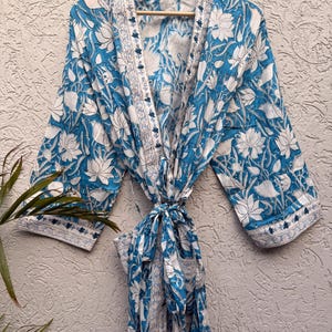 May include: A blue and white floral print robe hanging on a wooden hanger. The robe features a repeating pattern of white flowers on a blue background. The robe is tied at the waist with a matching fabric belt.