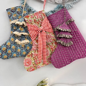 May include: Three festive Christmas stockings in various patterns. One is blue with floral motifs, another features a red and white striped bow, and the third is pink with a zig-zag design. A decorative garland hangs above.