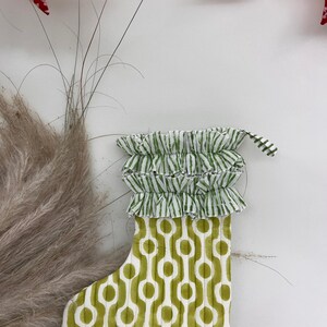 Op de afbeelding: Een decoratieve kerstsok met een groen en wit geometrisch patroon. De bovenkant van de sok heeft een gefranjerde rand met groen-witte strepen. Rode stervormige ornamenten hangen op de achtergrond. Een feestelijk item.