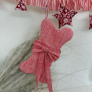 Puede incluir: Decoración navideña: un calcetín navideño a cuadros rojos y blancos con un lazo, colgado de una guirnalda a rayas a juego. La guirnalda presenta adornos de estrellas y triángulos.