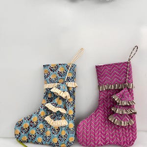 May include: Two decorative Christmas stockings. One is blue with a yellow floral pattern, the other is pink with a chevron pattern. Both have ruffled trim and hanging loops. The stockings are approximately 20 cm tall.