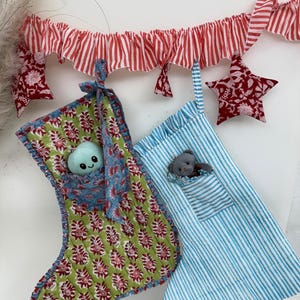 Puede incluir: Dos calcetines de Navidad, uno verde con estampado floral y otro azul con rayas blancas, cada uno con un pequeño animal de peluche. Una guirnalda a rayas rojas y blancas y adornos en forma de estrella cuelgan arriba.