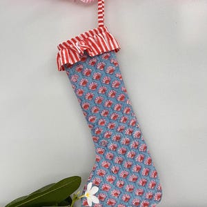 May include: A quilted Christmas stocking with a blue floral pattern and red accents. The top of the stocking has a red and white striped ruffle. A matching striped loop is attached for hanging. A pink and white striped ruffle is at the top.