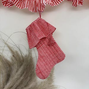May include: A red and white striped Christmas stocking hangs from a matching ruffled garland. The stocking and garland are made of fabric with a classic holiday pattern. The background is a plain white wall.