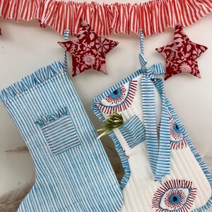 Puede incluir: Dos calcetines navideños, uno a rayas azules y blancas, el otro blanco con diseños de ojos rojos y azules. Adornos rojos en forma de estrella y una guirnalda a rayas rojas y blancas cuelgan arriba. Decoración festiva navideña.