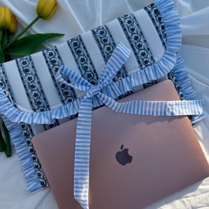 May include: A blue and white striped laptop sleeve with a ruffled edge and a blue and white striped ribbon tied in a bow. The sleeve is lying on a white surface with a laptop inside.
