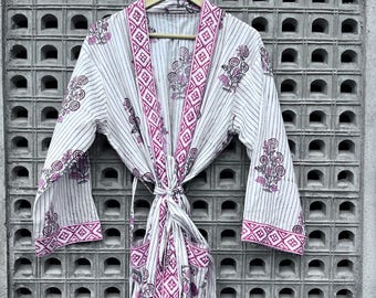 May include: A white robe with vertical stripes and floral and geometric patterns in shades of pink and purple. The robe has long sleeves and a tie closure at the waist. The robe is hanging on a wooden hanger.