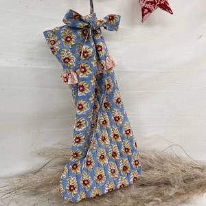 Op de afbeelding: Een blauwe kerstsok met een bloemenpatroon in rood en geel. De sok is gewatteerd en heeft een gerimpelde rand. Een bijpassende strik en ophanglus zijn bevestigd. Een rood-wit gestreepte slinger en een rood-wit patroonvlag bevinden zich erboven.