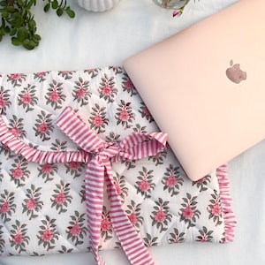 May include: A white quilted laptop sleeve with a pink floral design and a pink striped ribbon tie. A pink laptop sits beside the sleeve. The sleeve has a frilled edge. A small white vase and a small glass vase with pink flowers are also present.