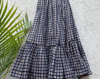 May include: A long, flowing skirt with a blue and tan checked pattern. The skirt is hanging on a wooden hanger against a textured white wall. The skirt has a tiered design with a ruffled hemline.
