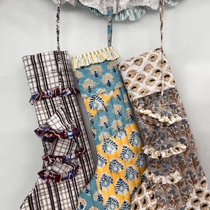 Puede incluir: Tres calcetines navideños acolchados con volantes y lazos para colgar. Un calcetín tiene un estampado de cuadros, otro tiene un diseño floral azul y amarillo, y el tercero tiene un estampado floral marrón y blanco. Una guirnalda decorativa cuelga encima.