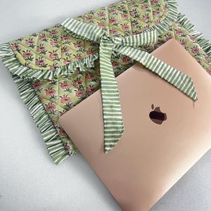 Puede incluir: Una funda acolchada para portátil en tela verde claro con un estampado floral rosa, con un borde con volantes y un cierre de lazo de tela a rayas verdes y blancas, colocada junto a un portátil color oro rosa con el logo de Apple visible.