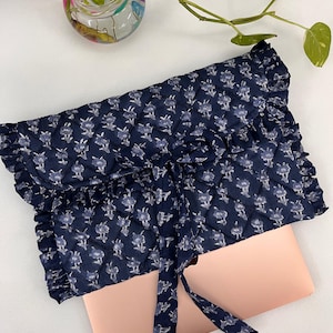 Pode incluir: Uma capa para laptop azul marinho com um padrão floral e bordas com babados. A capa é amarrada com uma fita de tecido combinando. Um laptop rosa é parcialmente visível. Uma tigela de vidro com pedras coloridas e uma planta estão ao fundo.