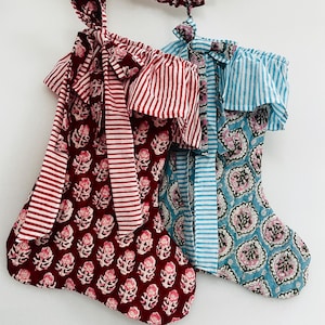 May include: Two Christmas stockings with floral and striped patterns. One is burgundy with white floral designs and red and white striped accents. The other is light blue with pink floral designs and blue and white striped accents. A burgundy garland hangs above.