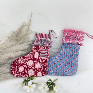 Peut inclure: Deux bas de Noël décoratifs. L'un est bordeaux avec un motif floral blanc, l'autre est bleu avec un motif floral rose. Les deux ont des volants et des boucles de suspension. Des fleurs blanches sont à la base des bas.