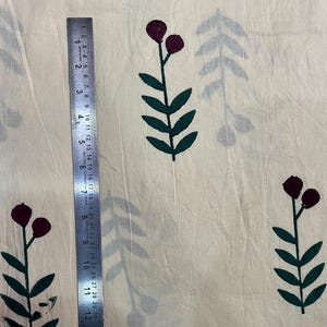 May include: Cream-colored fabric with a repeating pattern of dark green leaves and maroon berry-like shapes. A metal ruler, marked in inches, is placed vertically on the fabric, showing measurements up to 12 inches.
