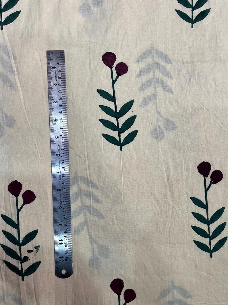 May include: Cream-colored fabric with a repeating pattern of dark green leaves and maroon berry-like shapes. A metal ruler, marked in inches, is placed vertically on the fabric, showing measurements up to 12 inches.