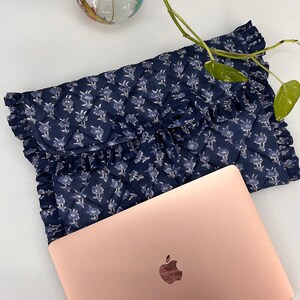 May include: A navy blue quilted laptop sleeve featuring a white floral pattern and ruffled edges, paired with a rose gold laptop. The sleeve has a fold-over flap design with a diamond-stitched texture and a soft, fabric construction.