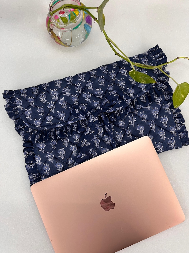 May include: A navy blue quilted laptop sleeve featuring a white floral pattern and ruffled edges, paired with a rose gold laptop. The sleeve has a fold-over flap design with a diamond-stitched texture and a soft, fabric construction.