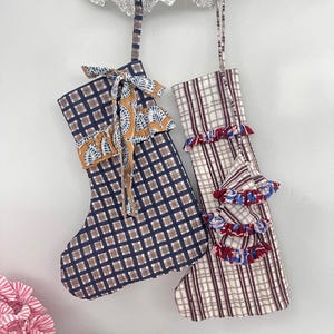 Peut inclure: Deux bas de Noël suspendus sur fond blanc. L'un est bleu marine avec un motif quadrillé marron et un nœud décoratif. L'autre est de couleur crème avec un motif à carreaux rouge et marron et une garniture à volants.