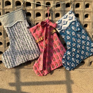 Peut inclure: Trois chaussettes de Noël en tissu suspendues à une ficelle. Les chaussettes sont décorées de motifs floraux différents. Une chaussette est bleue et blanche, une autre est rose et rouge, et la dernière est grise et blanche.