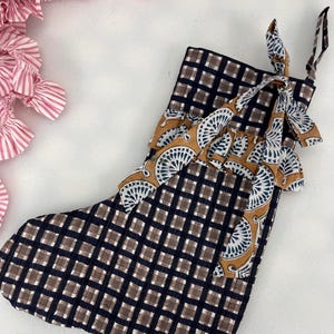 Könnte beinhalten: Ein Weihnachtsstrumpf mit einem marineblauen und braunen Karomuster. Der obere Rand hat ein dekoratives Band mit einem fächerartigen Design in Beige und Weiß. Der Strumpf hat eine Schlaufe zum Aufhängen. Rosa und weiß gestreifter Stoff ist im Hintergrund zu sehen.