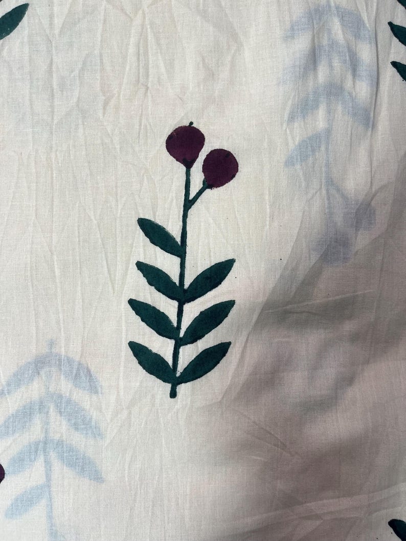 May include: White fabric with a repeating pattern of stylized botanical elements. The design features dark purple berries and dark green leaves on stems, with light blue leaf shapes scattered across the surface. The fabric has a slightly wrinkled texture.