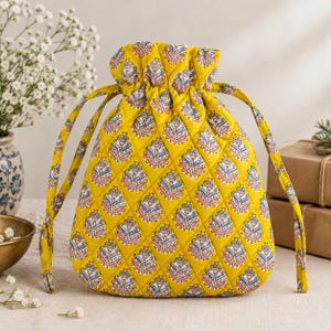 May include: A quilted drawstring pouch in a vibrant yellow fabric featuring a repeating blue and white floral block print pattern. The bag has a gathered top closure with two matching yellow fabric drawstrings on either side.