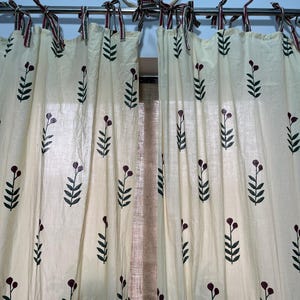 Op de afbeelding: Een paar crèmekleurige gordijnen met een groene en bordeauxrode bloemenprint. De gordijnen zijn vastgebonden met rode en groene gestreepte stofbanden.