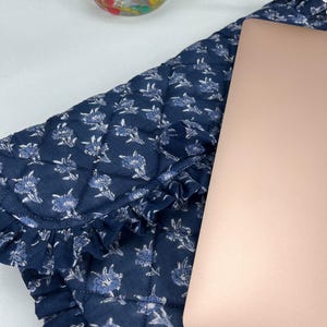 May include: A quilted navy blue laptop sleeve featuring a repeating light blue floral pattern and a decorative ruffled edge. The sleeve is partially covered by a rose gold laptop lid, showcasing a soft fabric texture and a structured design.