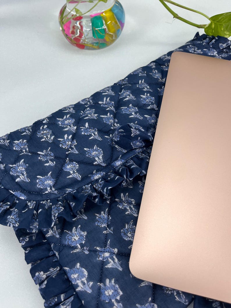 May include: A quilted navy blue laptop sleeve featuring a repeating light blue floral pattern and a decorative ruffled edge. The sleeve is partially covered by a rose gold laptop lid, showcasing a soft fabric texture and a structured design.