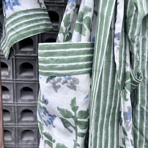 K&ouml;nnte beinhalten: Nahaufnahme eines wei&szlig;en Bademantels mit gr&uuml;nen und blauen Blumen- und Streifenmustern. Der Bademantel hat gr&uuml;ne Streifen an den &Auml;rmeln und Taschen. Der Stoff hat ein weiches, flie&szlig;endes Aussehen.
