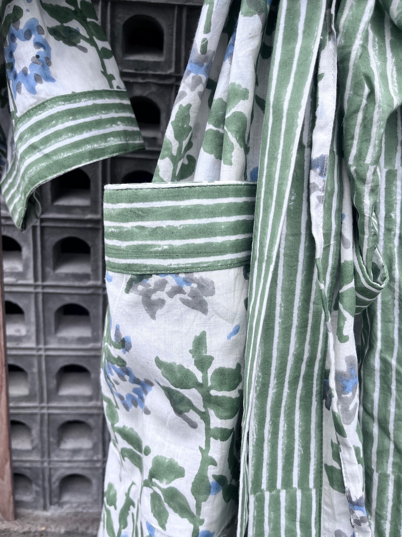 K&ouml;nnte beinhalten: Nahaufnahme eines wei&szlig;en Bademantels mit gr&uuml;nen und blauen Blumen- und Streifenmustern. Der Bademantel hat gr&uuml;ne Streifen an den &Auml;rmeln und Taschen. Der Stoff hat ein weiches, flie&szlig;endes Aussehen.