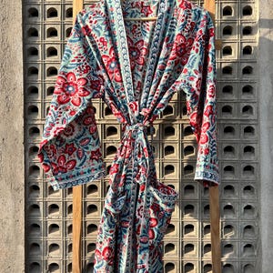 May include: A long, blue and red floral print robe with a tie belt and pockets. The robe is hanging on a wooden rack against a concrete wall.