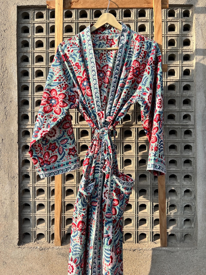 May include: A long, blue and red floral print robe with a tie belt and pockets. The robe is hanging on a wooden rack against a concrete wall.