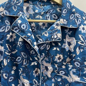 May include: A blue cotton shirt with a white floral print and a white trim around the collar. The shirt has a button-up closure.
