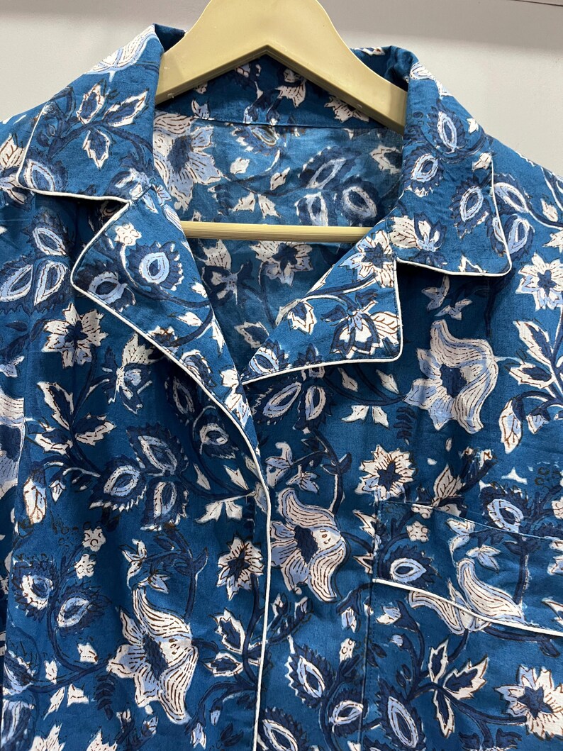 May include: A blue cotton shirt with a white floral print and a white trim around the collar. The shirt has a button-up closure.