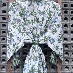 K&ouml;nnte beinhalten: Ein wei&szlig;es, floral gemustertes Gewand mit blauen und gr&uuml;nen Akzenten h&auml;ngt an einem Holzgestell. Das Gewand hat ein botanisches Design mit blauen Blumen und gr&uuml;nen Bl&auml;ttern. Der Kragen und die Manschetten haben einen gr&uuml;nen Streifen.