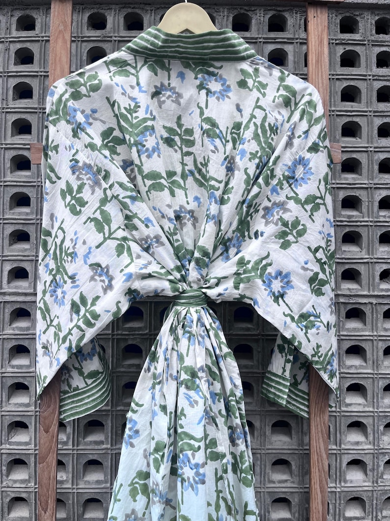 K&ouml;nnte beinhalten: Ein wei&szlig;es, floral gemustertes Gewand mit blauen und gr&uuml;nen Akzenten h&auml;ngt an einem Holzgestell. Das Gewand hat ein botanisches Design mit blauen Blumen und gr&uuml;nen Bl&auml;ttern. Der Kragen und die Manschetten haben einen gr&uuml;nen Streifen.