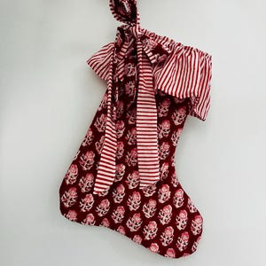 Puede incluir: Un calcetín navideño con un estampado floral en burdeos y blanco. La parte superior del calcetín tiene un estampado a rayas rojas y blancas, con un lazo a juego. Una guirnalda burdeos es visible en el fondo.