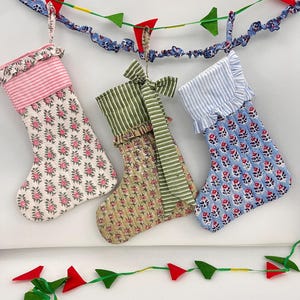 以下が含まれることがあります： 花柄とストライプ柄のクリスマスストッキング3つ。ストッキングはピンク、緑、青で、上部にフリルが付いています。赤と緑の三角旗のガーランドが上に吊り下げられています。