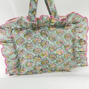 May include: A quilted tote bag with a floral pattern in shades of green, pink, and yellow. The bag has a ruffled edge with pink trim and matching handles. The bag is designed with a diamond pattern.