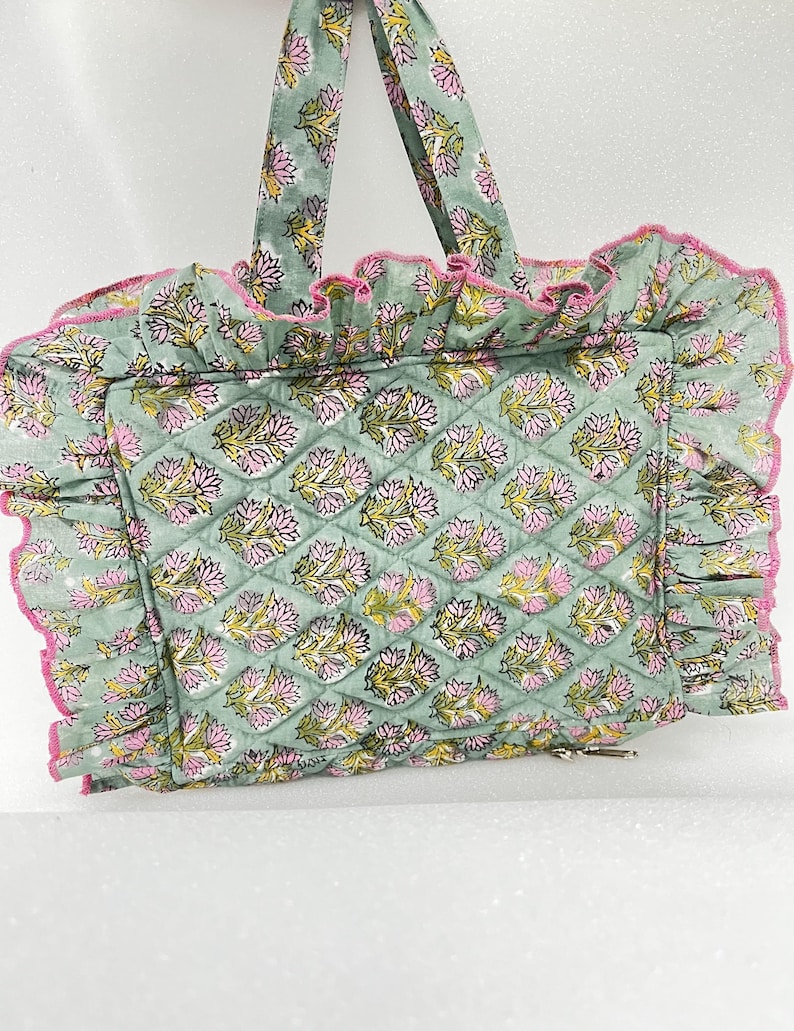 May include: A quilted tote bag with a floral pattern in shades of green, pink, and yellow. The bag has a ruffled edge with pink trim and matching handles. The bag is designed with a diamond pattern.