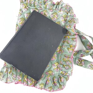 May include: A sage green book cover with a floral pattern, featuring pink and yellow flowers, rests on a matching quilted fabric with a ruffled edge. The fabric has ties and a pink trim.