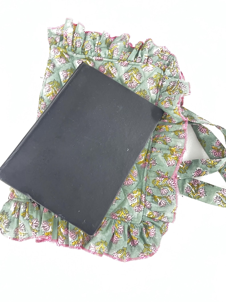 May include: A sage green book cover with a floral pattern, featuring pink and yellow flowers, rests on a matching quilted fabric with a ruffled edge. The fabric has ties and a pink trim.