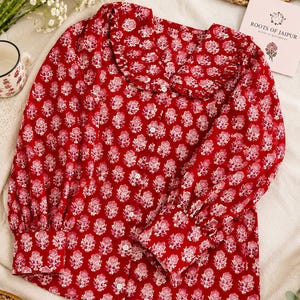 May include: A long-sleeved button-down shirt in a vibrant red color featuring a repeating white floral block print pattern. The shirt includes a wide, rounded collar with ruffled edges and buttoned cuffs, creating a structured yet soft appearance.