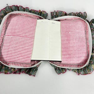 May include: An open notebook inside a floral print case with a pink and white striped lining. The case has a ruffled edge and a zip closure. The notebook is open to a blank page with the word "Notes" visible.